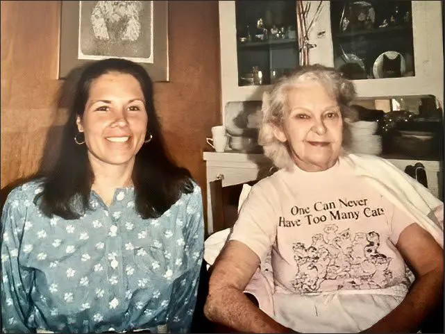 Two women smiling indoors, one younger with long hair, the other older wearing a cat-themed t-shirt.