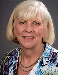 A woman with short blonde hair and a patterned top smiling against a gray background.