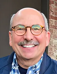 A smiling man with glasses and a mustache against a brick wall background.