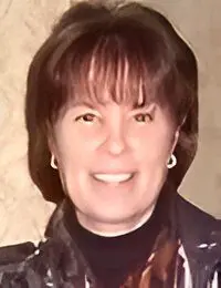 A smiling woman with short brown hair wearing a black top and earrings against a beige background.